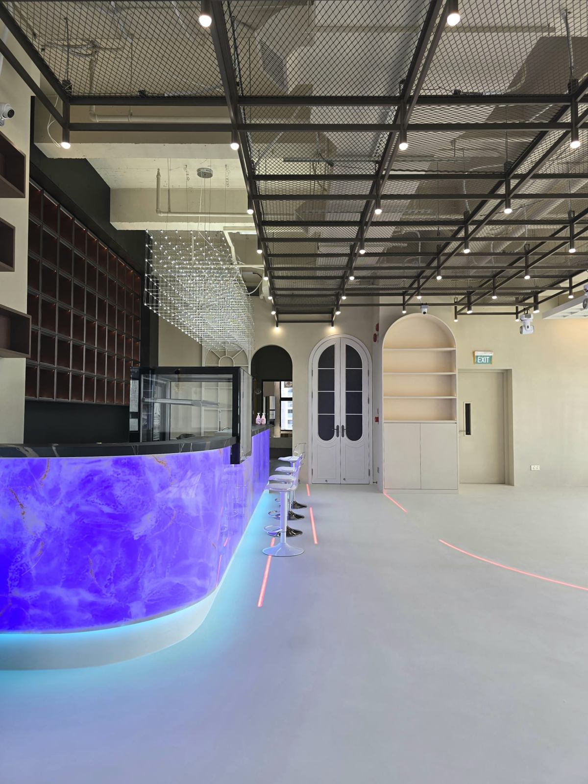 Open studio floor with a curved, backlit violet counter, clear acrylic stools, a black steel ceiling grid and white arched double doors.