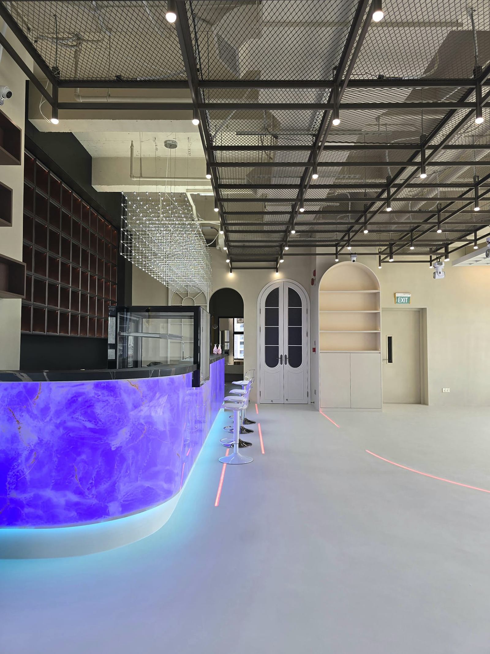 Open studio floor with a curved, backlit violet counter, clear acrylic stools, a black steel ceiling grid and white arched double doors.