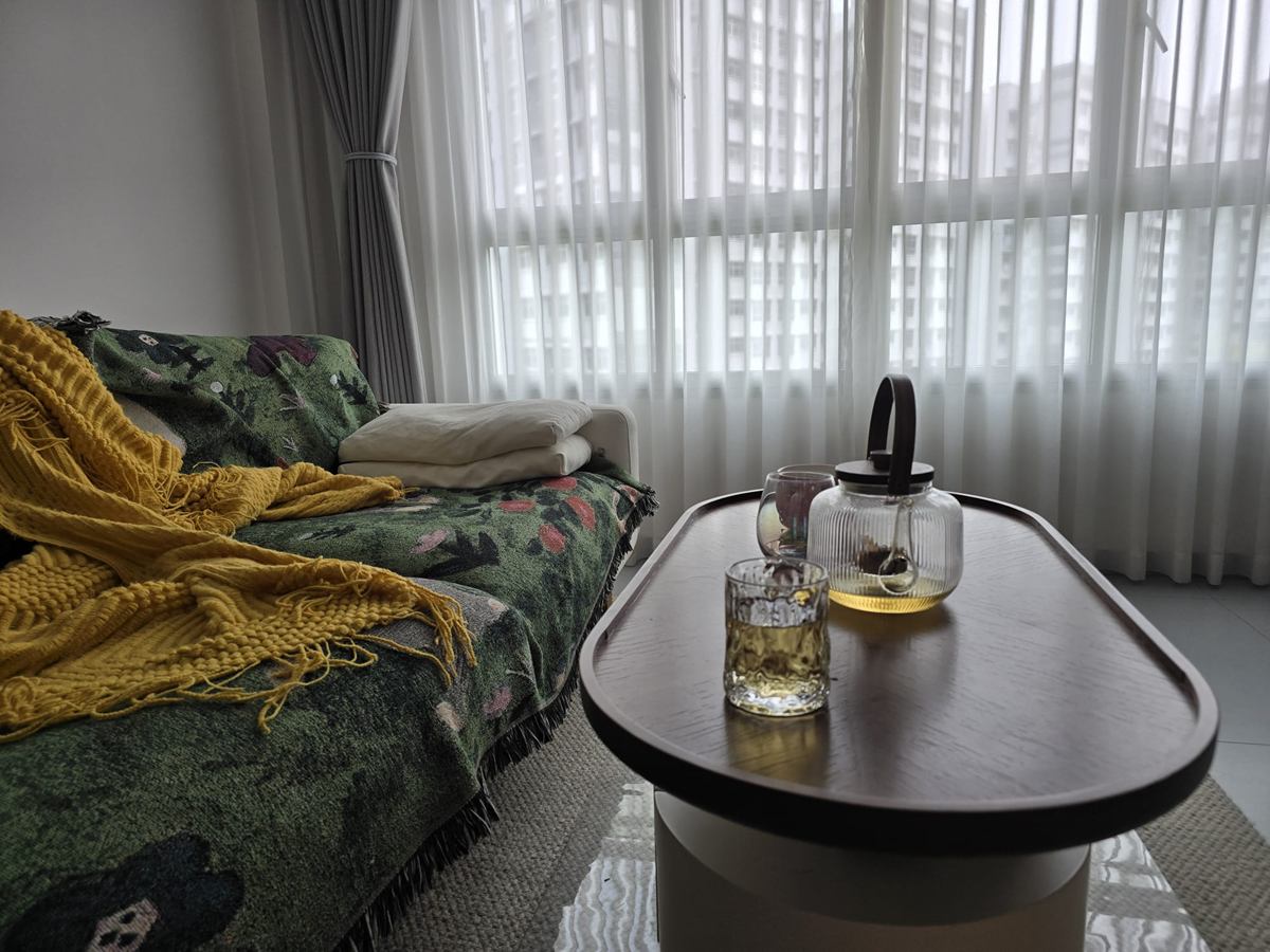 An oval timber coffee table with a glass teapot and cups beside a sofa with a green throw and yellow blanket, in front of sheer curtains.