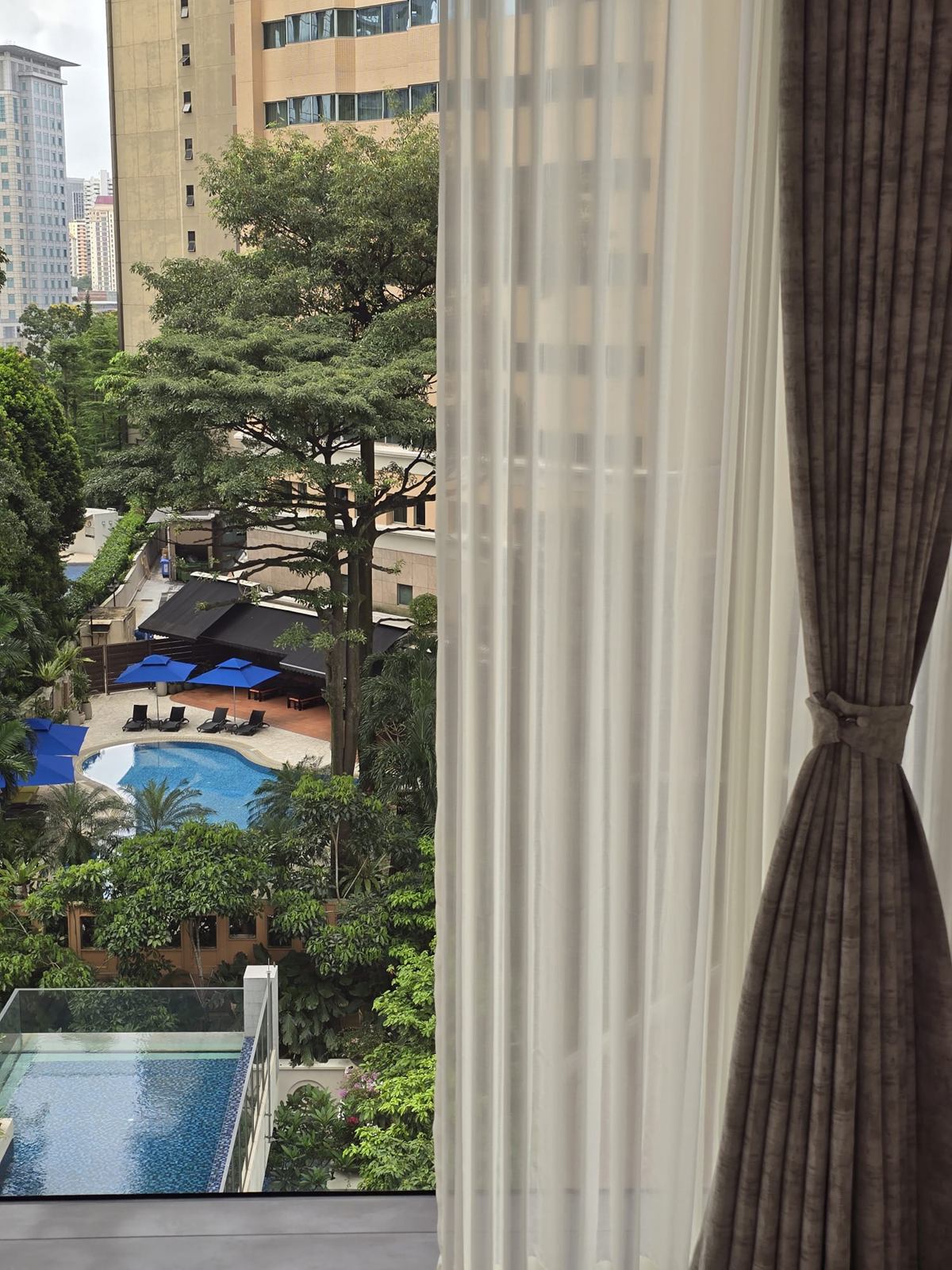 View from a window framed by sheer and dark curtains, over trees to a swimming pool below.