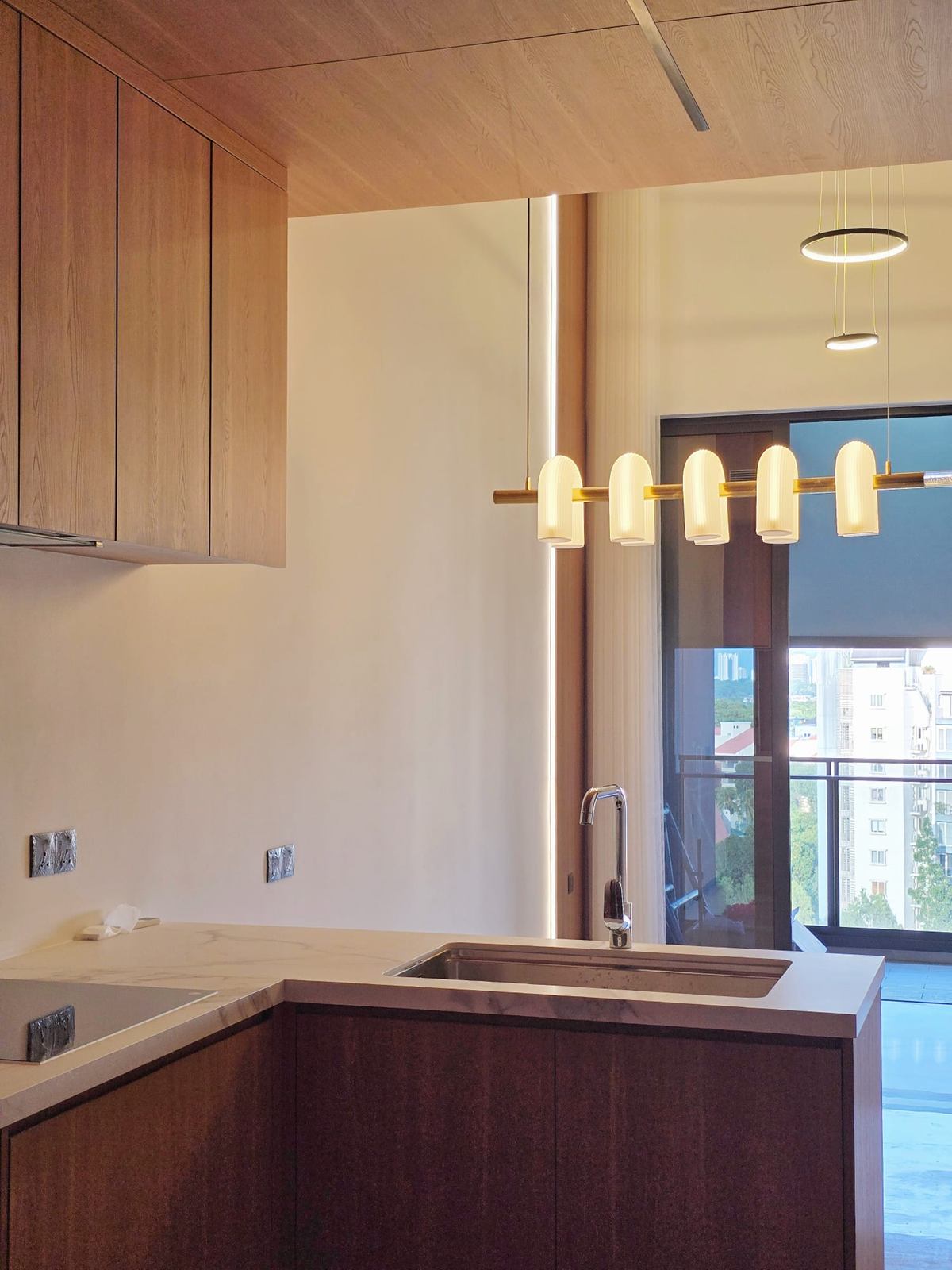 The kitchen seen towards the balcony door, with timber-toned cabinets, a brass pendant and a ring light beyond.